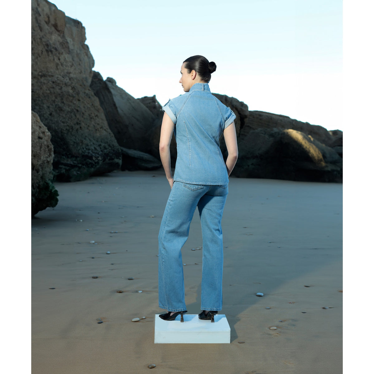 Wearing the La Chemise Cravate — Light Blue by SOEUR &amp; FRÈRES, a woman stands on a square platform on a sandy beach, her back to the camera as she faces large rocks in the distance. Crafted from 100% cotton and featuring an integrated tie detail, her relaxed denim shirt complements the tranquil coastal scenery.