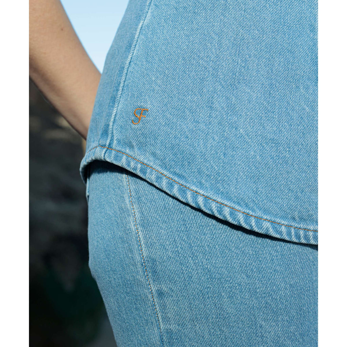 Close-up of a person wearing the SOEUR &amp; FRÈRES La Chemise Cravate — Light Blue, a relaxed, light blue 100% cotton denim shirt featuring a small gold “SF” logo on the top. The background is blurred.