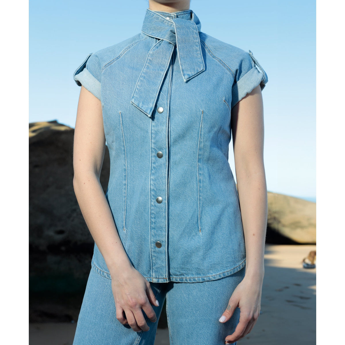 Wearing SOEUR &amp; FRÈRES' La Chemise Cravate in light blue—a relaxed-fit 100% cotton denim shirt with an integrated tie at the high neck—paired with matching denim pants, this person stands outdoors on a sandy beach with rocks in the background.
