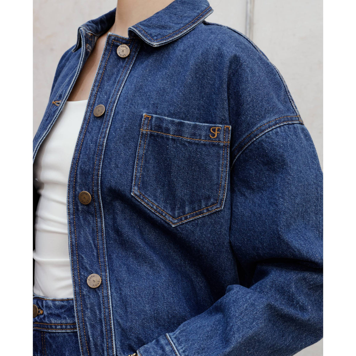 A person wears the SOEUR &amp; FRÈRES La Veste Courte — Stone Blue |PR Sample, a cropped denim jacket in a stone blue wash, over a white top. The jacket features decorative stitching on the chest pocket, metallic buttons, and a structured fit. The background is a plain, light-colored wall.