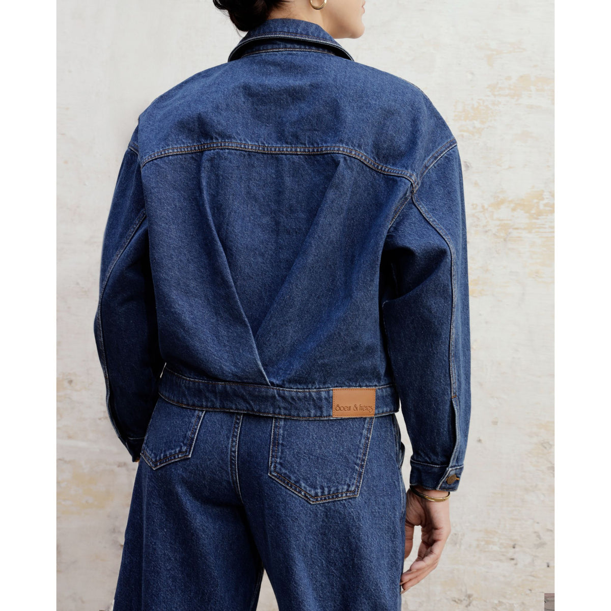 A person stands with their back to the camera against a light-colored textured wall, wearing the SOEUR &amp; FRÈRES La Veste Courte — Stone Blue |PR Sample, a cropped denim jacket and jeans in a structured fit.
