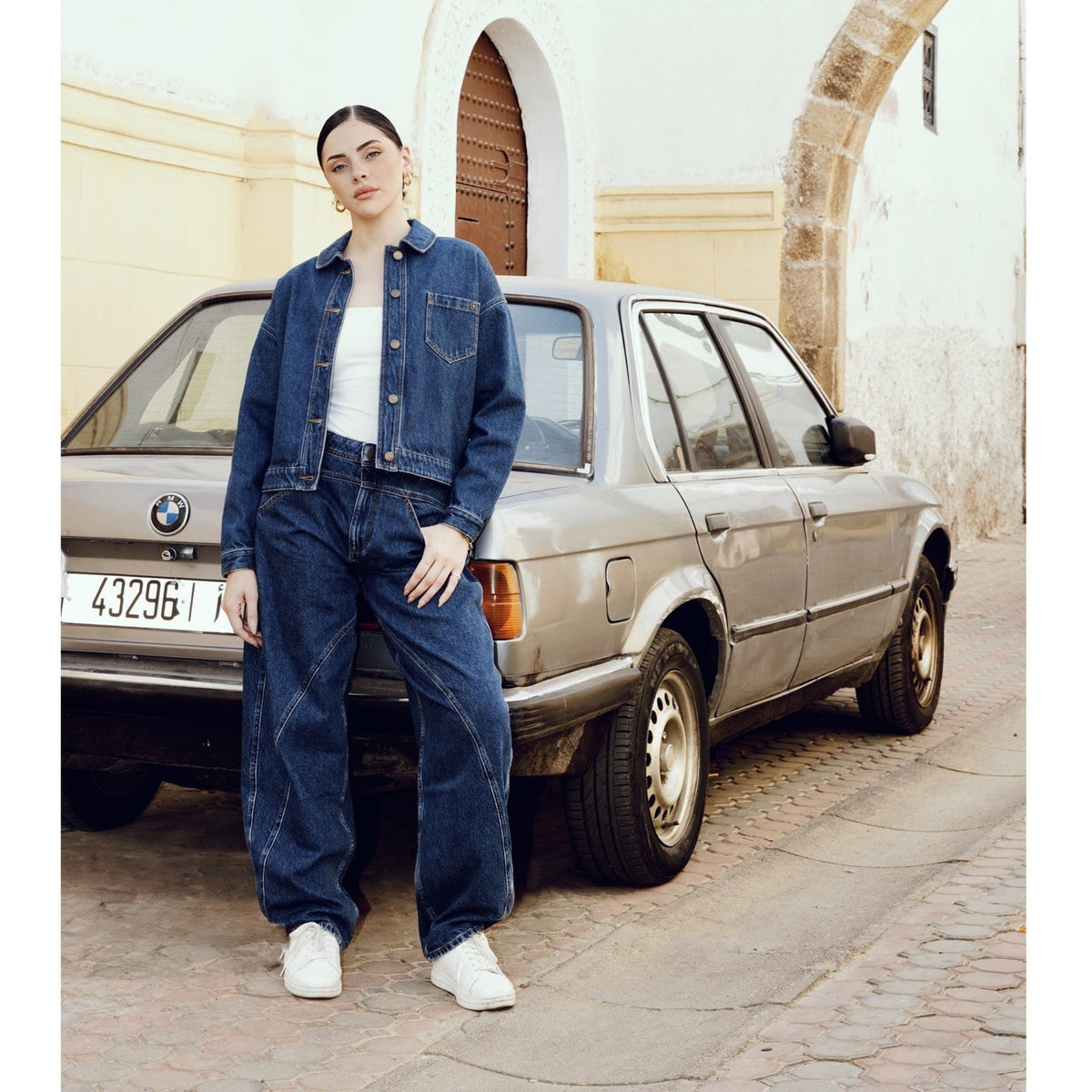 Wearing the SOEUR &amp; FRÈRES La Veste Courte in Stone Blue (PR Sample), a woman in a structured, cropped denim jacket and jeans leans against an old beige BMW parked on a cobblestone street near a white arched building.