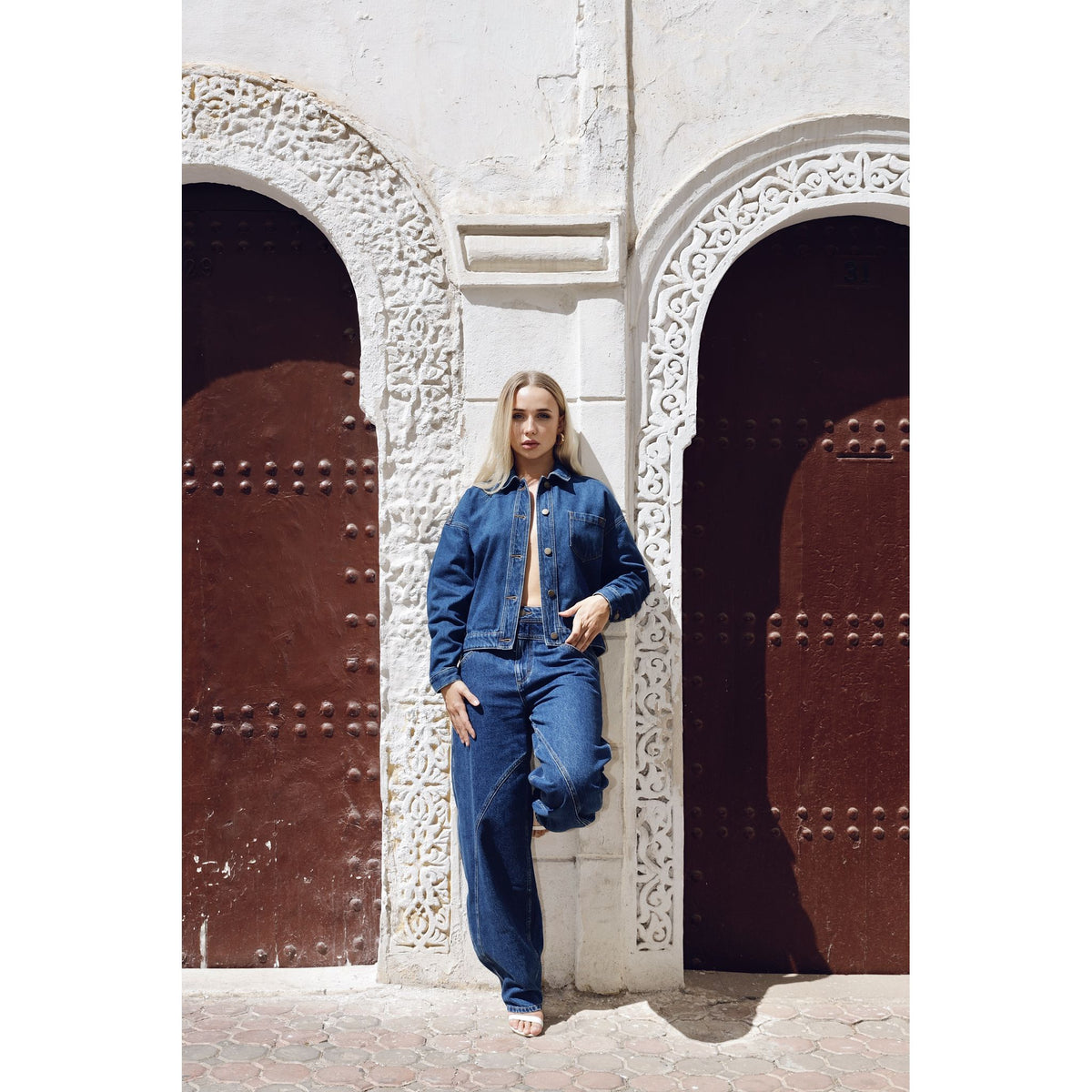 A woman stands confidently between two ornate, dark wooden doors set in a white, textured wall, wearing the SOEUR &amp; FRÈRES La Veste Courte — Stone Blue |PR Sample, a cropped denim jacket featuring a structured fit and stone blue wash.