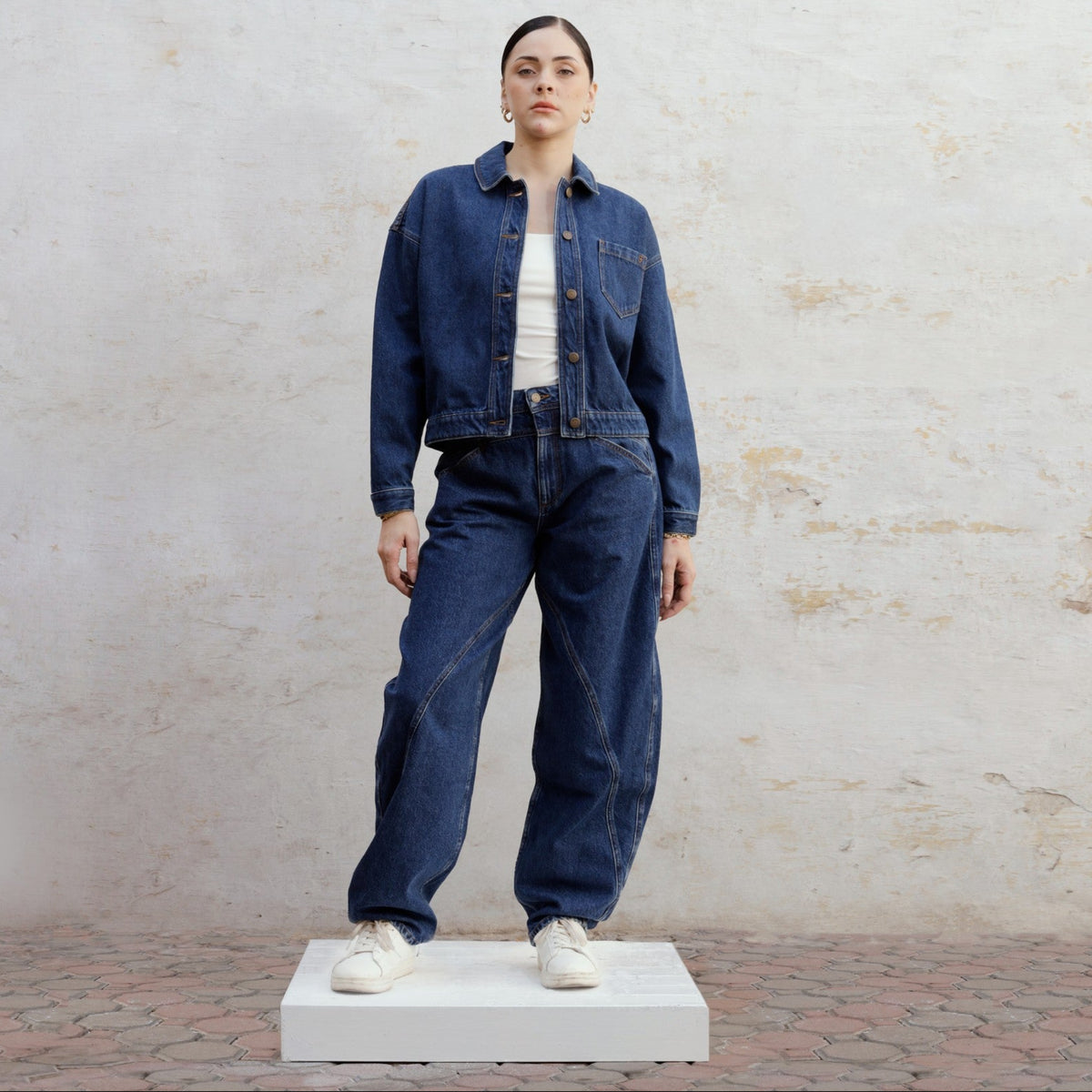 A person stands on a white platform, wearing SOEUR &amp; FRÈRES' relaxed fit denim jacket and Le Barrel — Stone Blue jeans (PR Sample), styled with a white shirt and sneakers, against a light textured wall and brick ground.