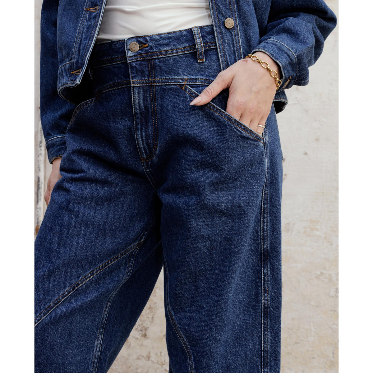 Wearing the SOEUR &amp; FRÈRES Le Barrel — Stone Blue (PR Sample) high-waisted, relaxed fit barrel-leg jeans and a denim jacket, this person stands against a light-colored wall with one hand in their pocket, accessorized with a gold bracelet and ring on their left hand.