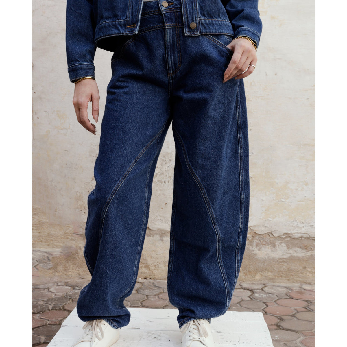 A person is wearing SOEUR &amp; FRÈRES' Le Barrel — Stone Blue |PR Sample jeans, styled with a matching denim jacket and white sneakers. They are standing outdoors on a textured surface against a neutral wall background, with only the lower half of their body visible.