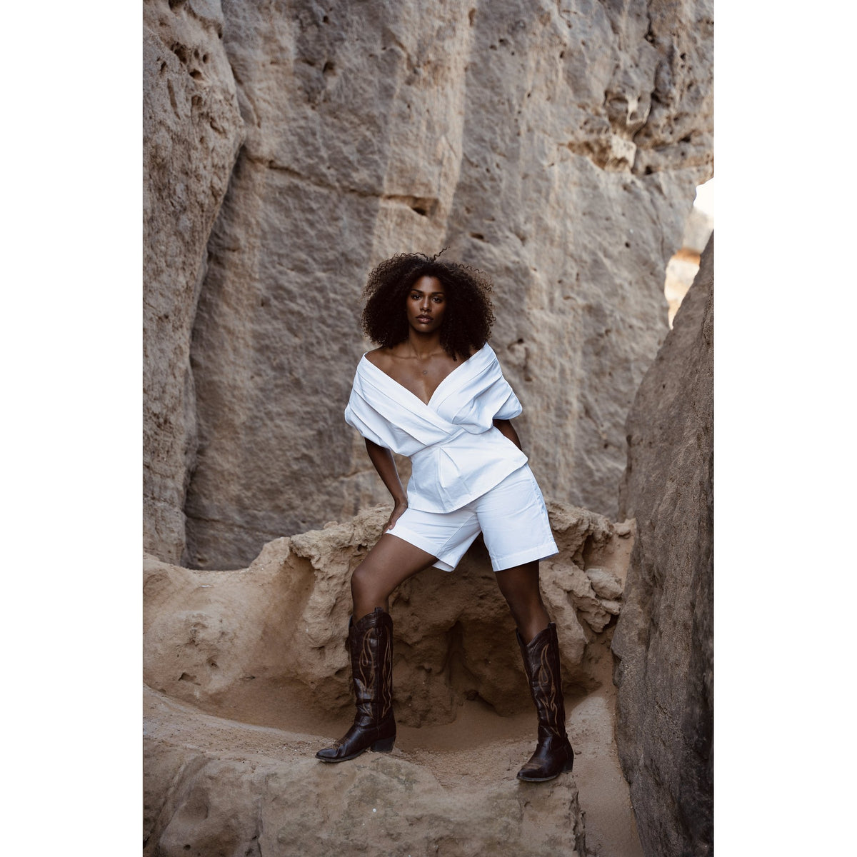 A woman with curly hair poses confidently among large rock formations, wearing the Le Bermuda — Optic White by SOEUR &amp; FRÈRES, styled with a chic white off-shoulder top and brown cowboy boots.