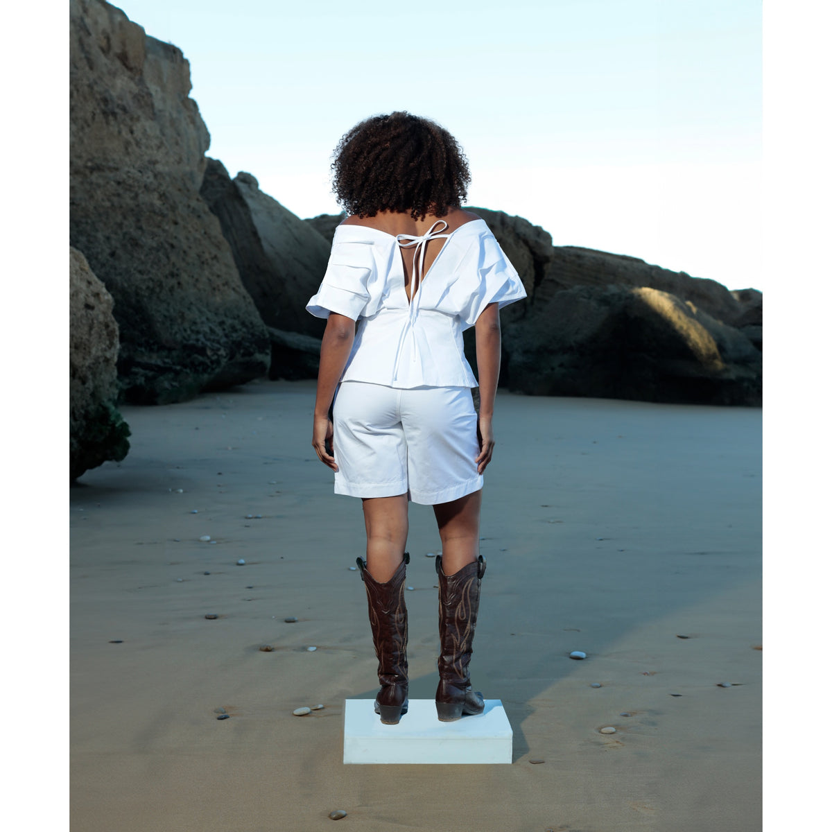 A person with curly hair stands on a beach platform, facing away. They wear the Le Off Shoulder — Optic White top by SOEUR &amp; FRÈRES, styled with matching white denim shorts and dark cowboy boots for a true statement look. Large rocks and sand appear in the background.