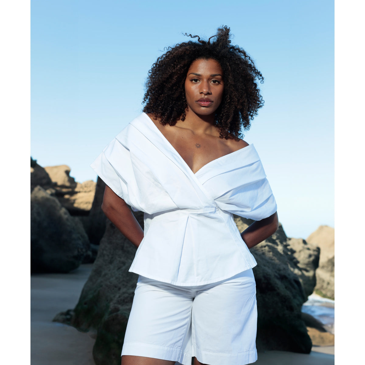 Certainly! Here’s a rewritten version of the sentence, incorporating your product data: A woman with curly hair stands on a rocky beach, wearing SOEUR &amp; FRÈRES’ Le Off Shoulder in Optic White—a chic PR Sample—paired with sleek white denim shorts, set against the backdrop of a clear blue sky.