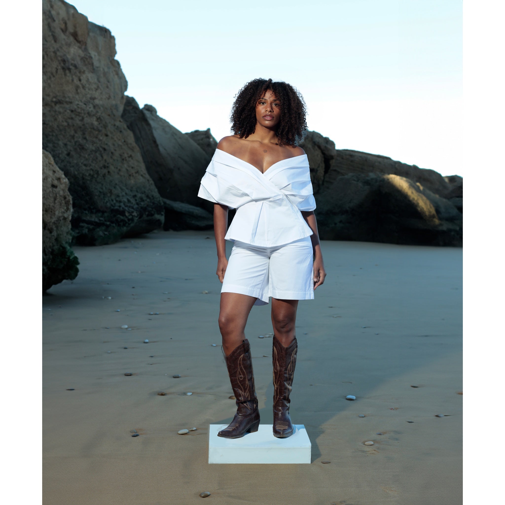 A woman stands on a small platform on a sandy beach, wearing SOEUR & FRÈRES' Le Off Shoulder top in Optic White (PR Sample) and shorts with brown cowboy boots, large rocks in the background.