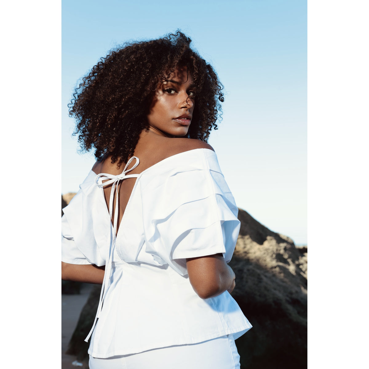 A woman with curly hair wears the SOEUR &amp; FRÈRES Le Off Shoulder top in Optic White—a true statement piece featuring a bow at the back—as she stands outdoors against a rocky background under a clear blue sky.