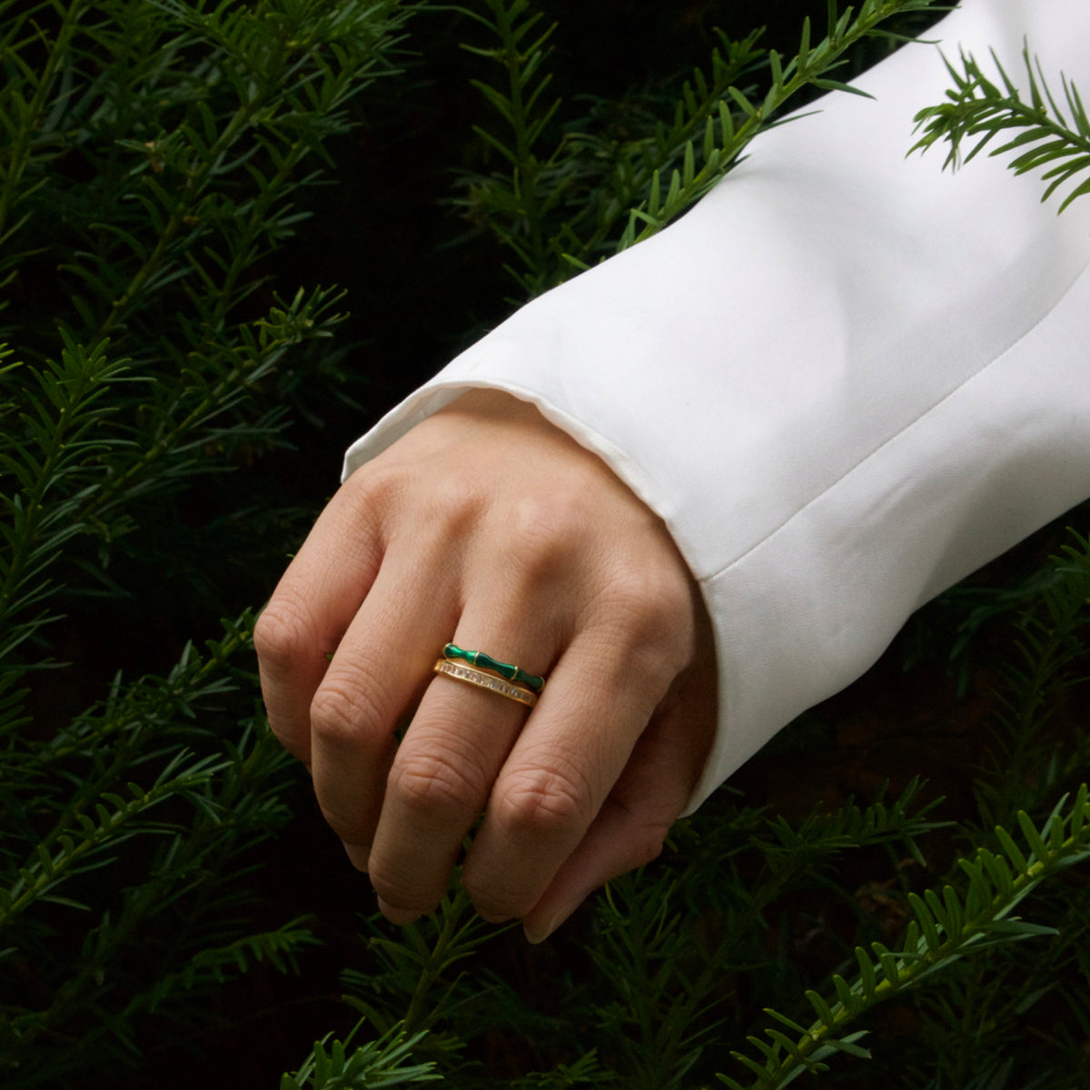 Hand wearing a gold ring with a green stone on a background of green foliage