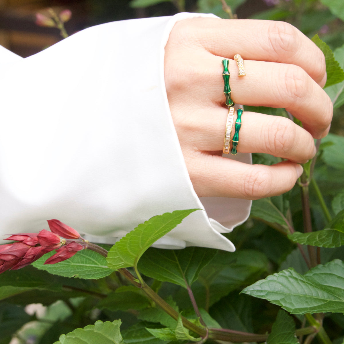 Hand wearing a gold ring with green gemstones against a natural background