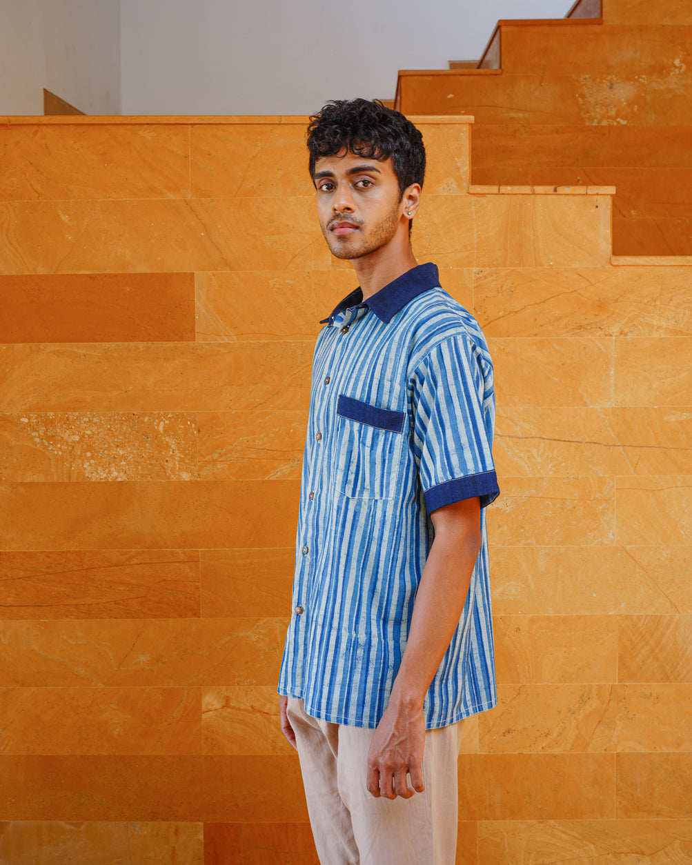 A young man with short curly hair stands sideways in front of an orange stone staircase, wearing JIWYA's Neel Striped Shirt in blue with indigo signature elements and cream pants.