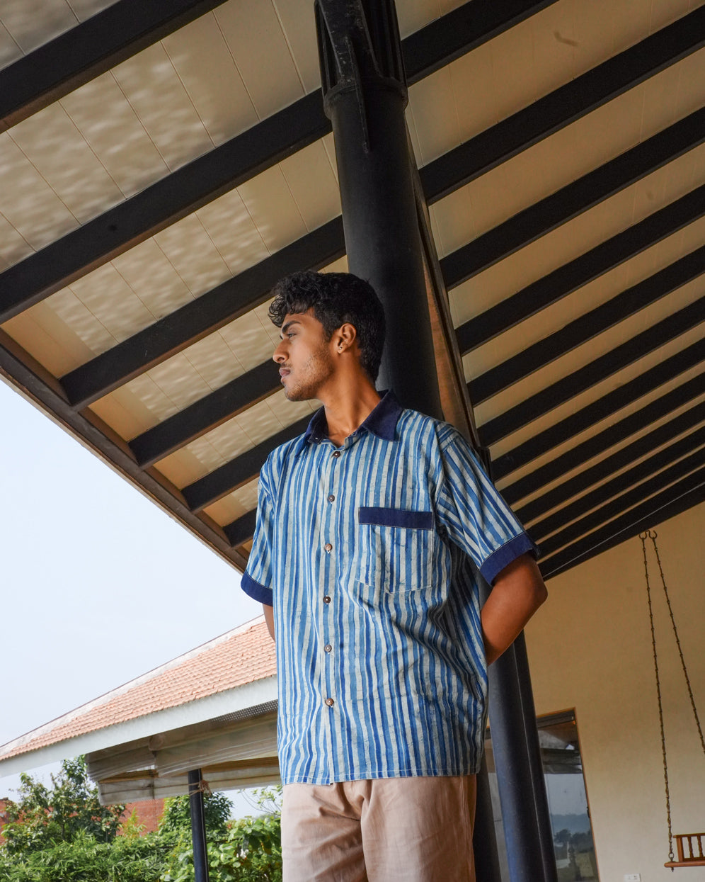 A young man stands under a covered outdoor area, wearing the JIWYA Neel Striped Shirt in blue with indigo signature elements. He looks to the left with hands behind his back; in the background are a building, trees, and a partially cloudy sky.