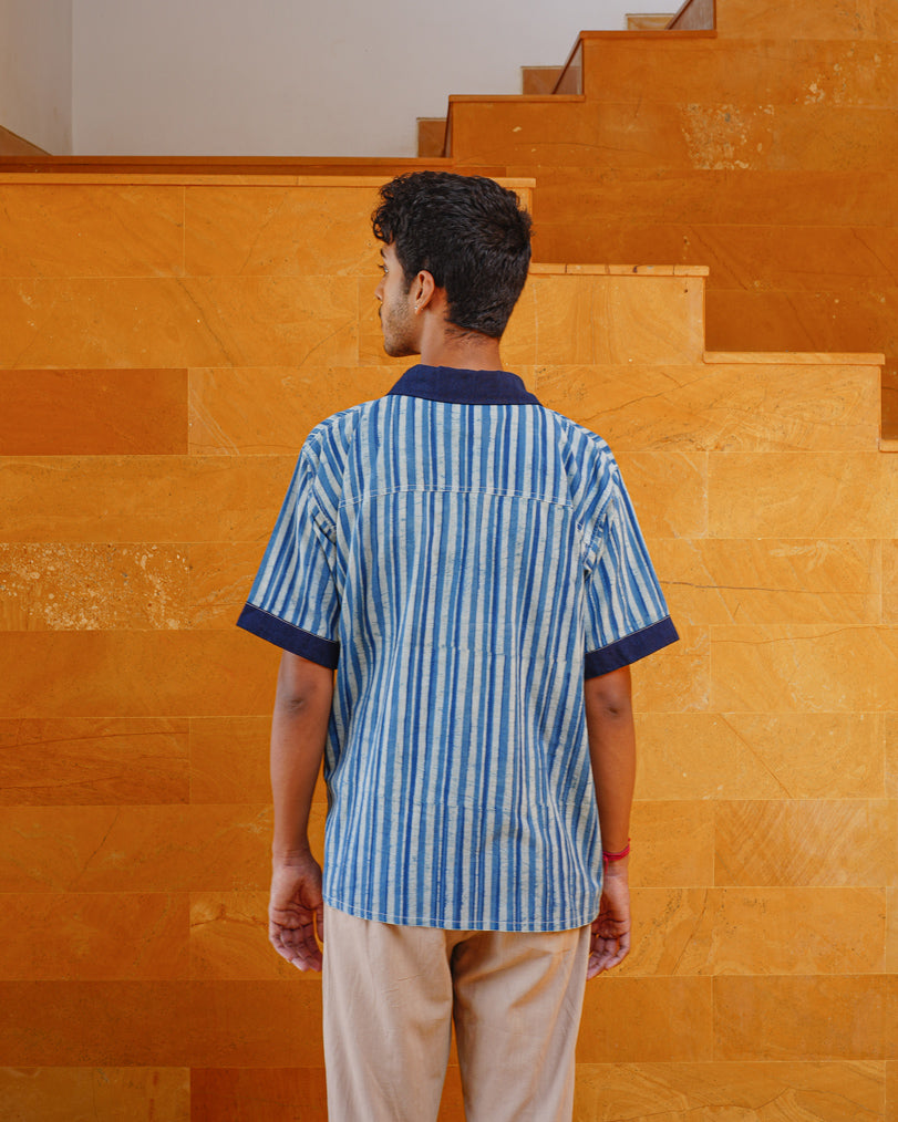 A person stands facing away, wearing JIWYA's Neel Striped Shirt in blue with indigo signature elements, paired with light beige pants. They are in front of a yellow-orange tiled staircase and wall.