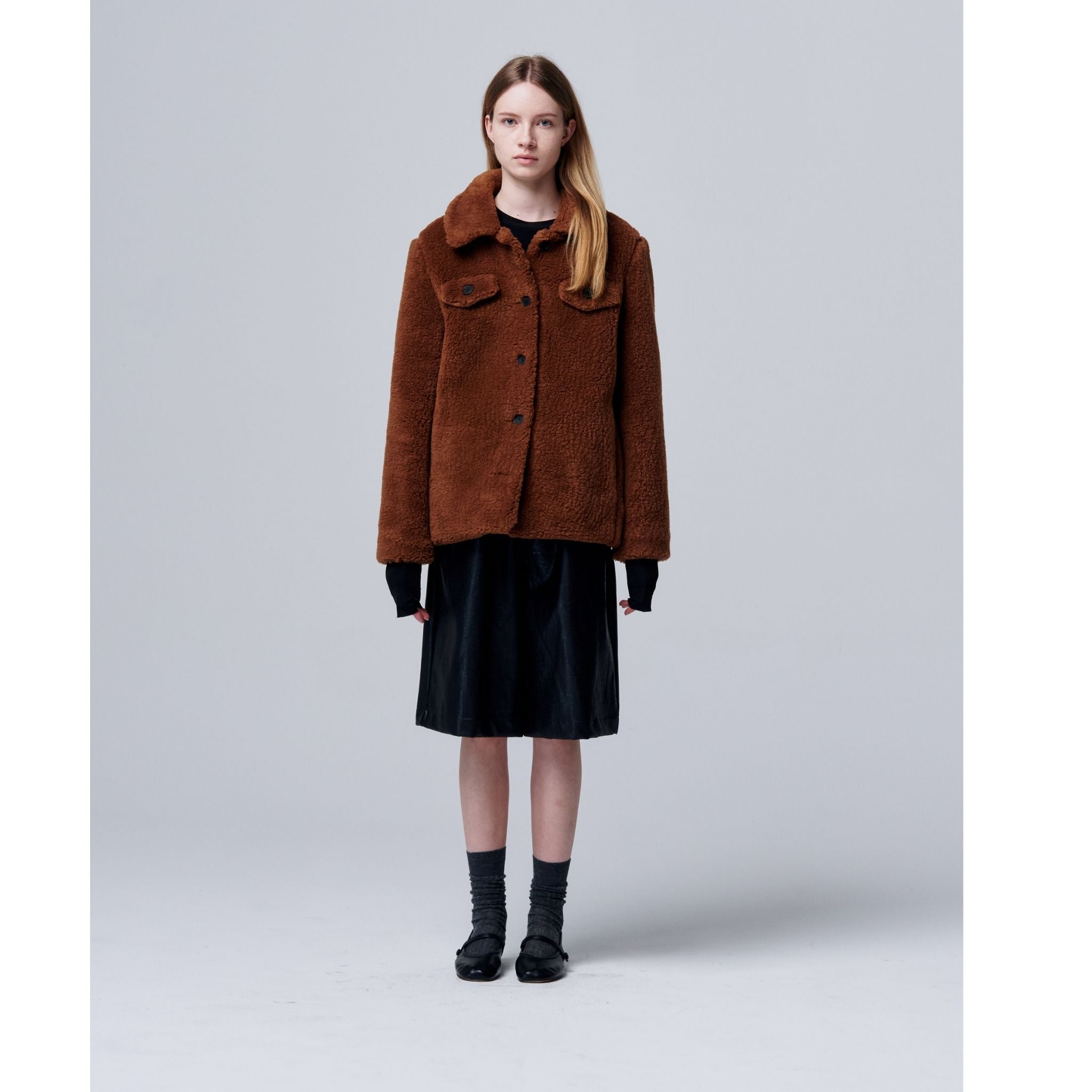 A young woman with long, straight hair and a neutral expression stands against a gray background, wearing the Ethereal Fur Jacket with Soft Fleece Fabric over a black dress, styled with gray socks and black shoes—an easy winter layering piece.