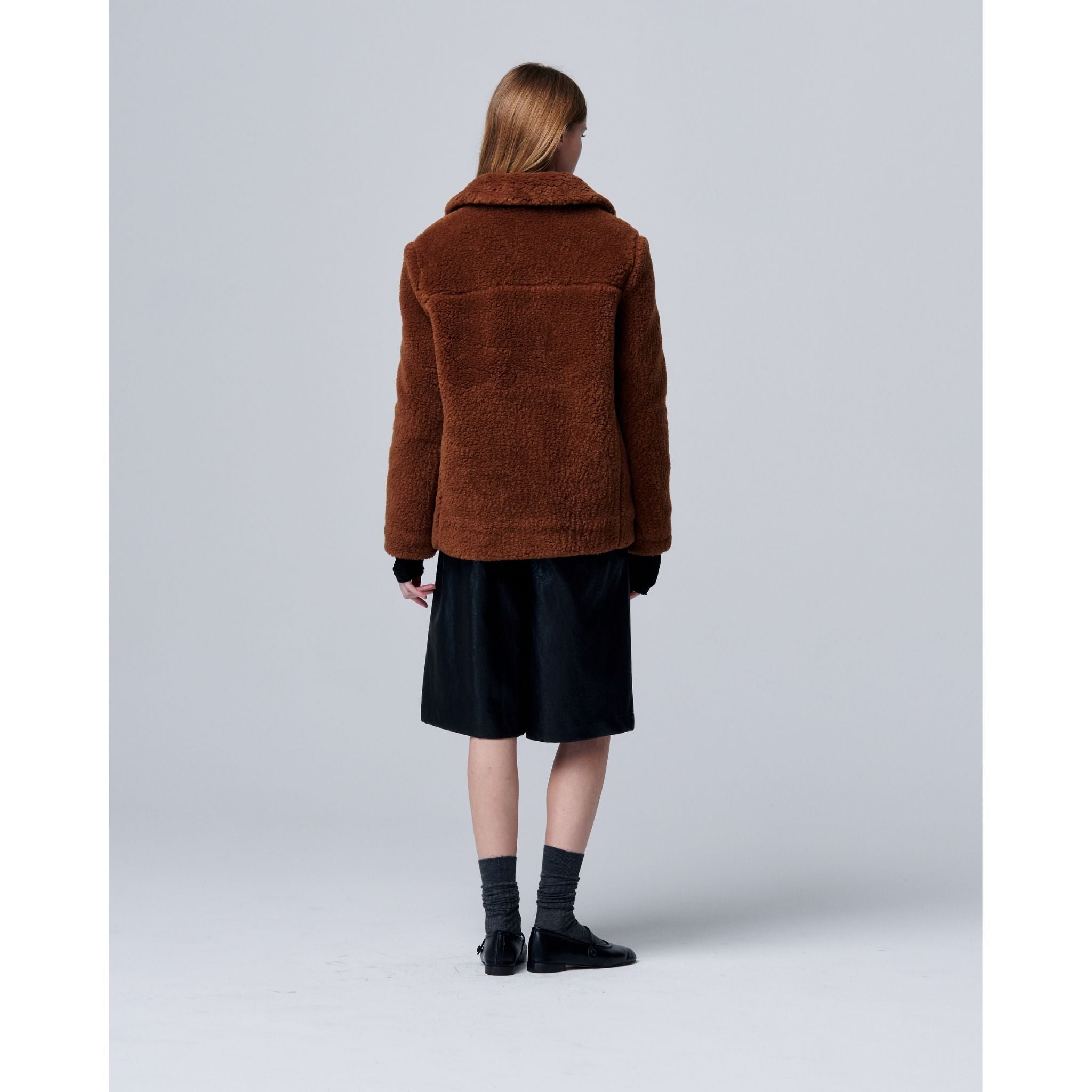 A person with straight, long hair stands facing away, wearing the Ethereal Fur Jacket with Soft Fleece Fabric as a cozy winter layer, paired with black knee-length shorts, gray socks, and black shoes against a plain light background.