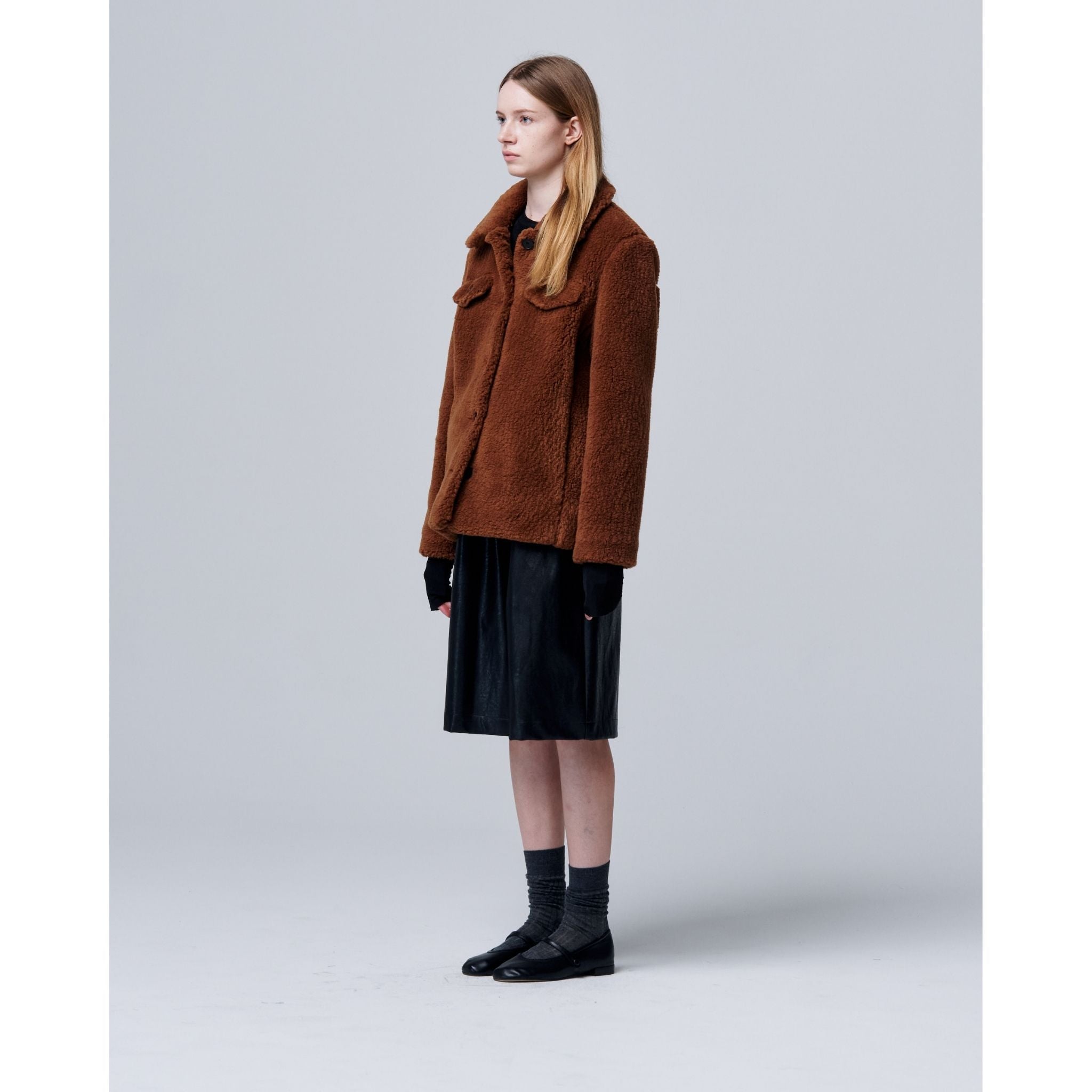 A person with long, straight hair stands in profile against a gray background, wearing the Ethereal Fur Jacket with Soft Fleece Fabric as a winter layer, paired with a black skirt, gloves, socks, and black shoes.