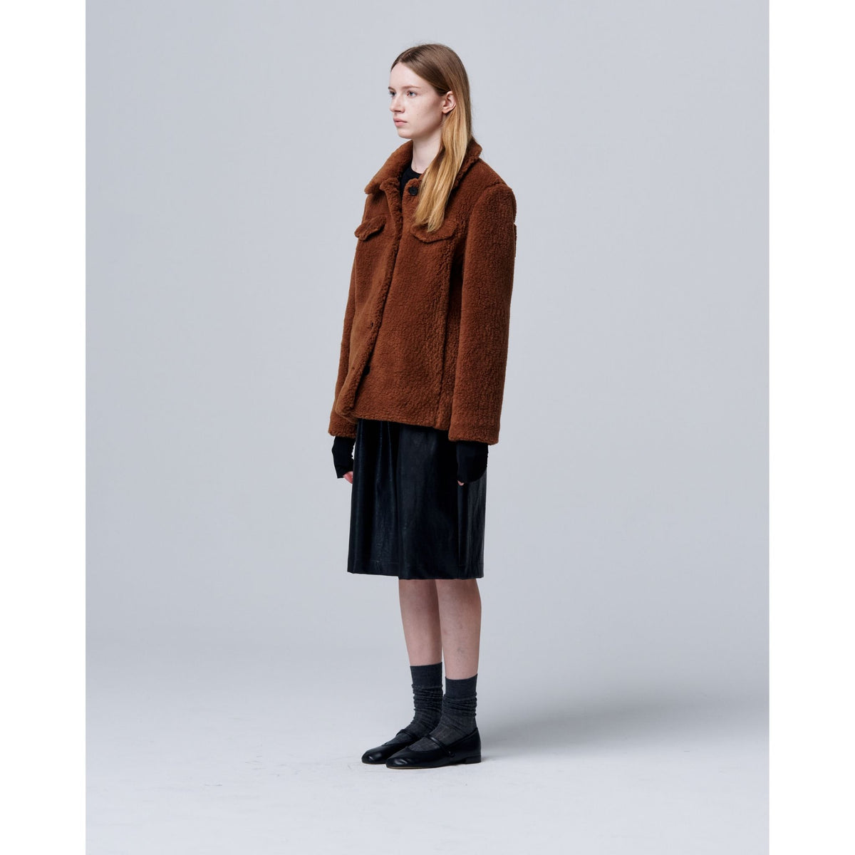 A person with long, straight hair stands in profile against a gray background, wearing the Ethereal Fur Jacket with Soft Fleece Fabric as a winter layer, paired with a black skirt, gloves, socks, and black shoes.