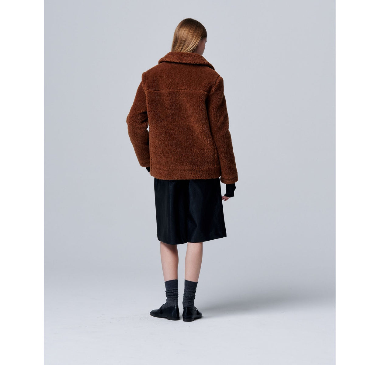 A person with straight, shoulder-length hair, seen from behind, models the Ethereal Fur Jacket with Soft Fleece Fabric over a black knee-length skirt, black shoes, and gray socks against a plain light gray background.