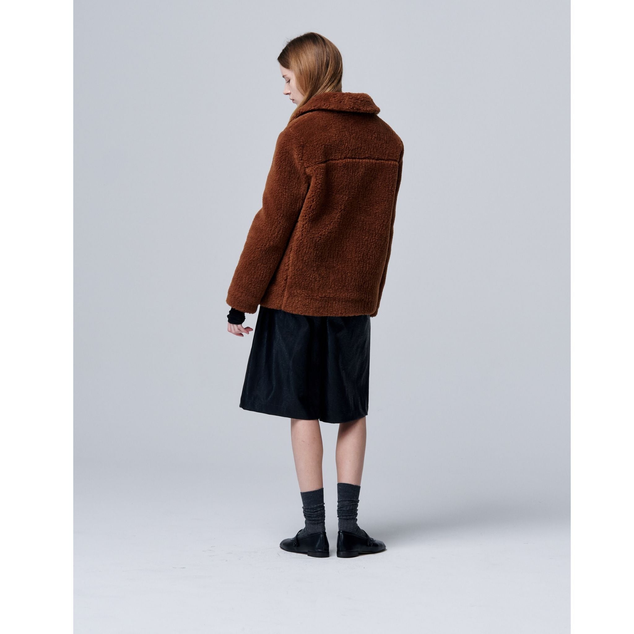 A person stands facing away, wearing the Ethereal Fur Jacket with Soft Fleece Fabric, a black knee-length skirt, dark socks, and black shoes against a plain light gray background.