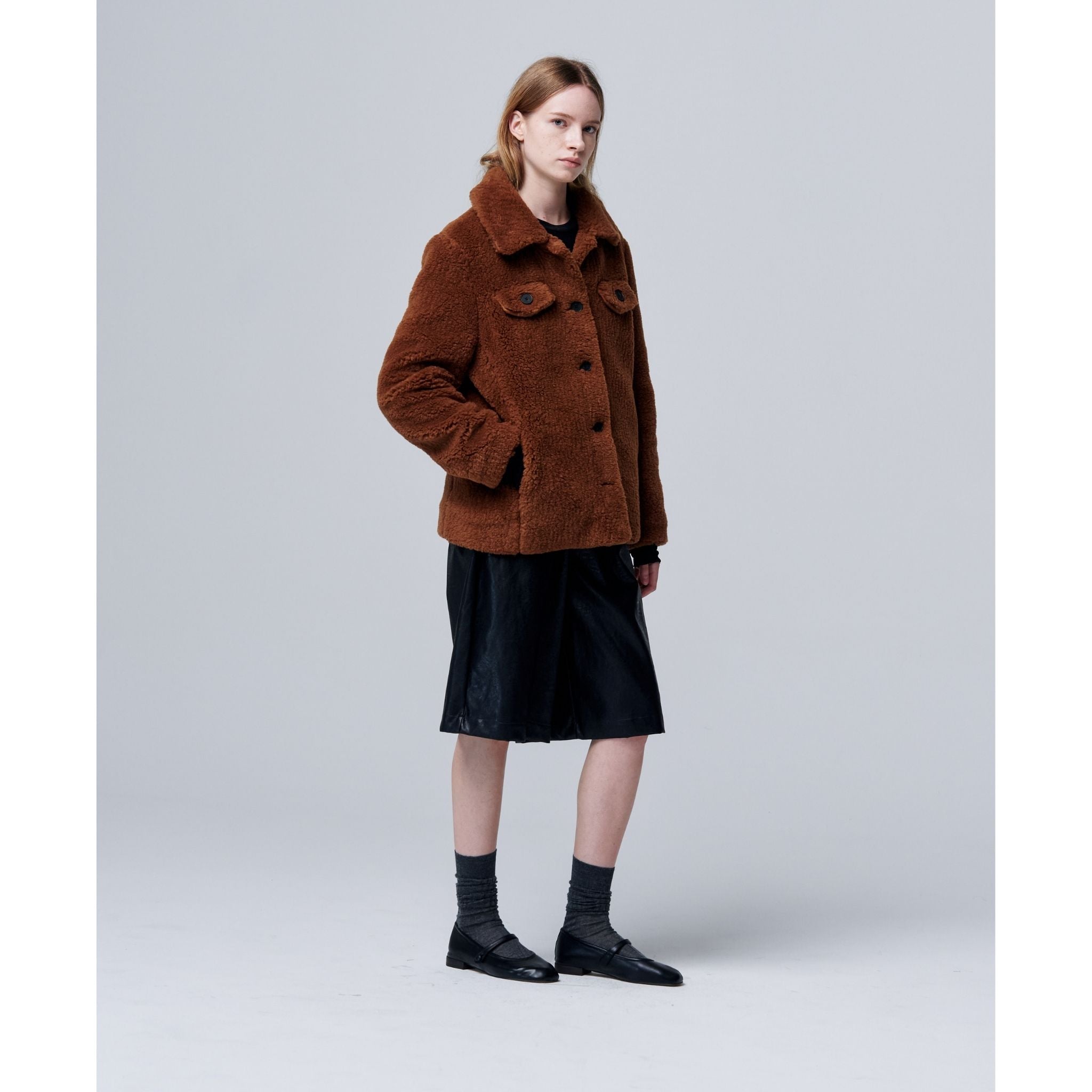 A person with light brown hair stands against a plain background, wearing an Ethereal Fur Jacket with Soft Fleece Fabric as a winter layer, paired with a black knee-length skirt, black socks and shoes, one hand in their jacket pocket.