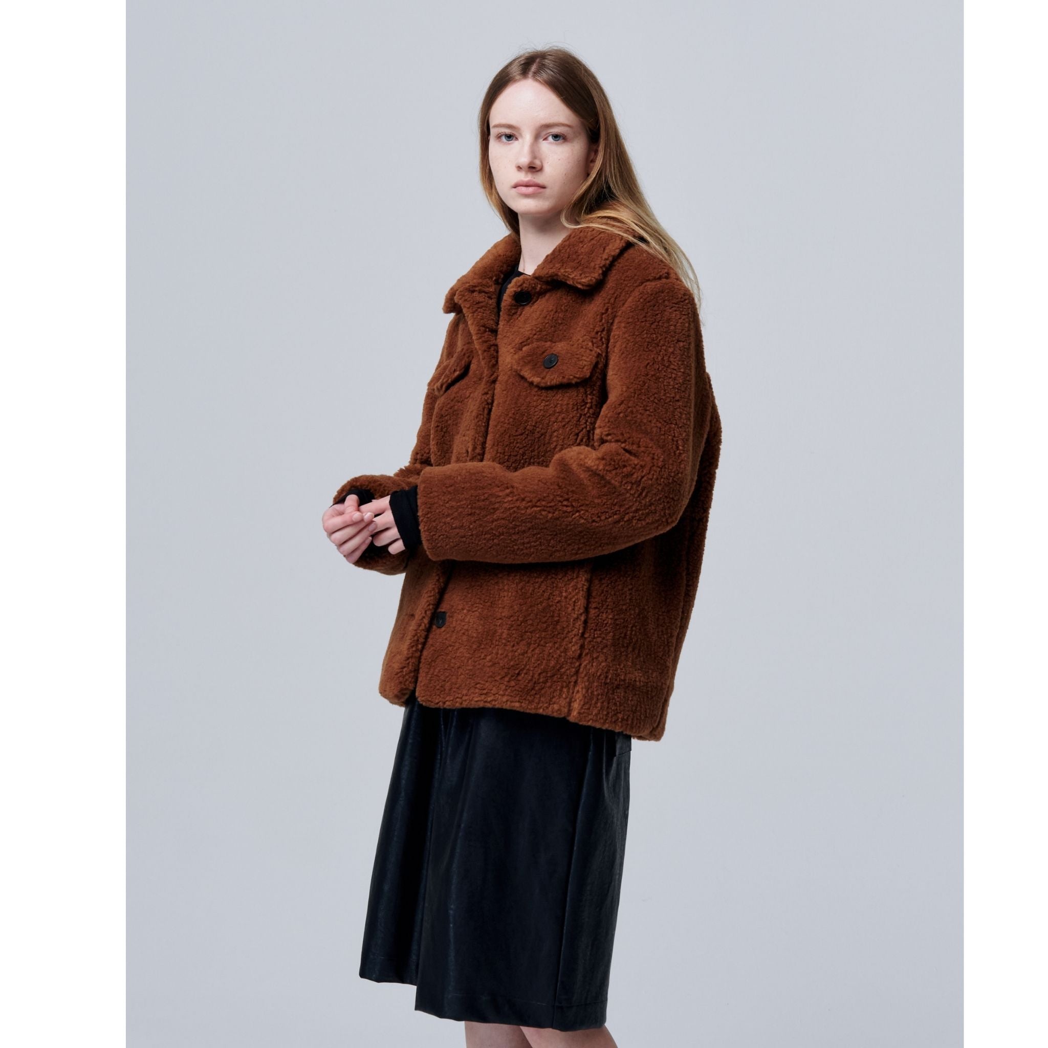 A woman with long, straight hair wears the Ethereal Fur Jacket with Soft Fleece Fabric in brown, featuring textured material and buttons, paired with a black skirt as she stands against a plain light gray background.