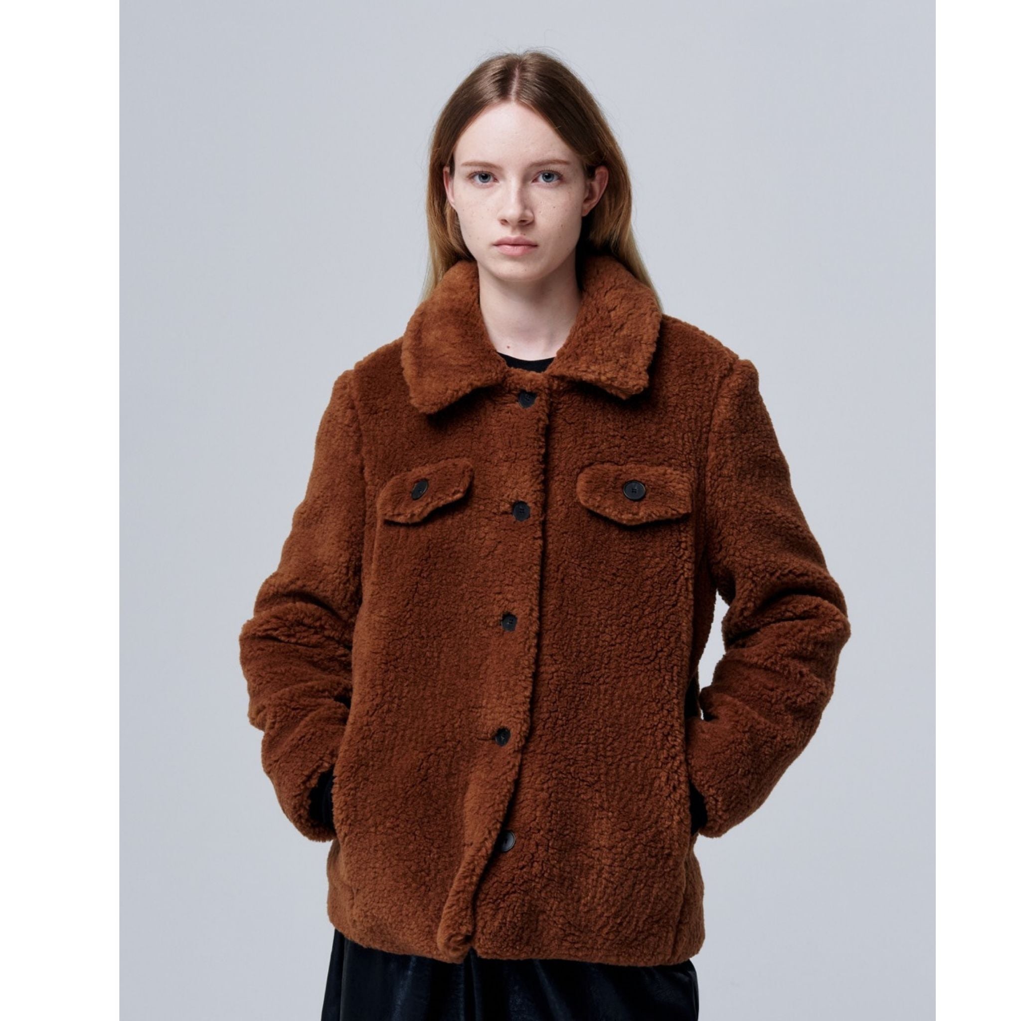 A person with straight, light brown hair models the Ethereal Fur Jacket—a cozy, brown sherpa-style button-up with front pockets—against a light gray background. This soft fleece piece is perfect for winter layering.