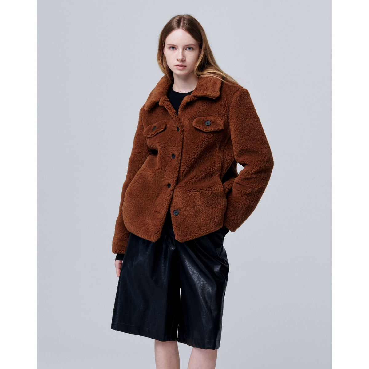 A woman with straight, light brown hair wears the Ethereal Fur Jacket with Soft Fleece Fabric over a black top and knee-length black leather shorts, standing against a plain light gray background—a perfect winter layering piece.