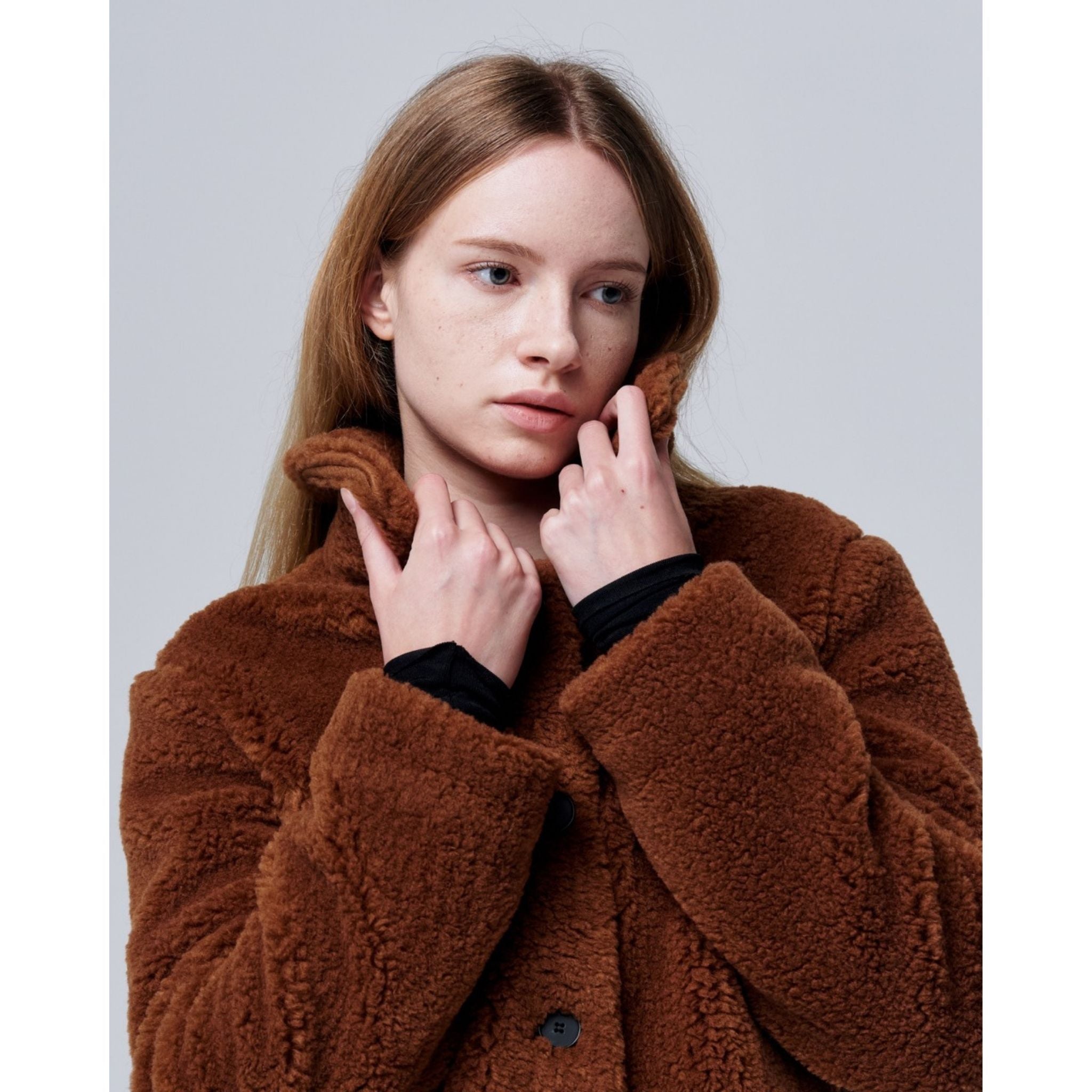 A young woman with long blonde hair models the Ethereal Fur Jacket with Soft Fleece Fabric, gently holding the collar with both hands as she looks to the side against a plain light background.