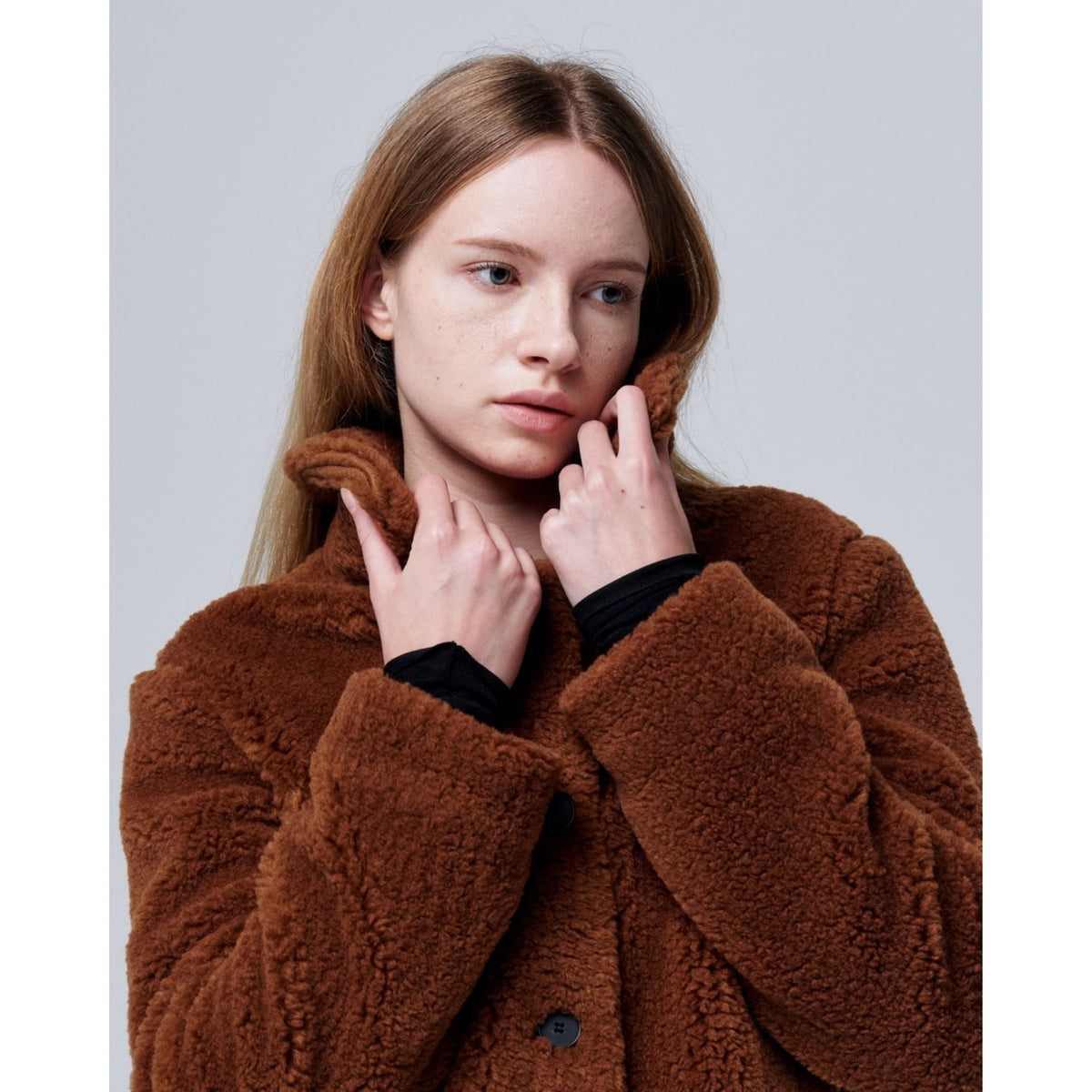 A young woman with long blonde hair models the Ethereal Fur Jacket with Soft Fleece Fabric, gently holding the collar with both hands as she looks to the side against a plain light background.
