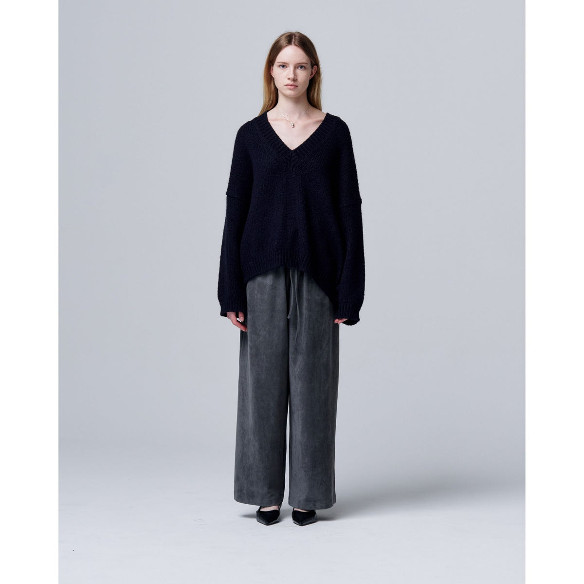 A woman stands against a plain gray background, wearing a loose black V-neck sweater, ODDPRESENT Velour Suede Pants with Elastic Waistband, and black shoes. She faces forward neutrally, arms relaxed at her sides.
