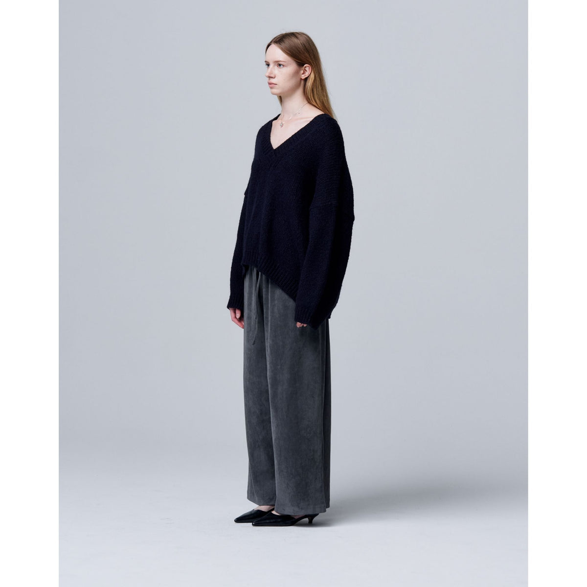 A woman with straight, shoulder-length hair and a neutral expression stands in profile against a plain gray background, wearing ODDPRESENT Velour Suede Pants with Elastic Waistband, a dark loose sweater, and black heels.