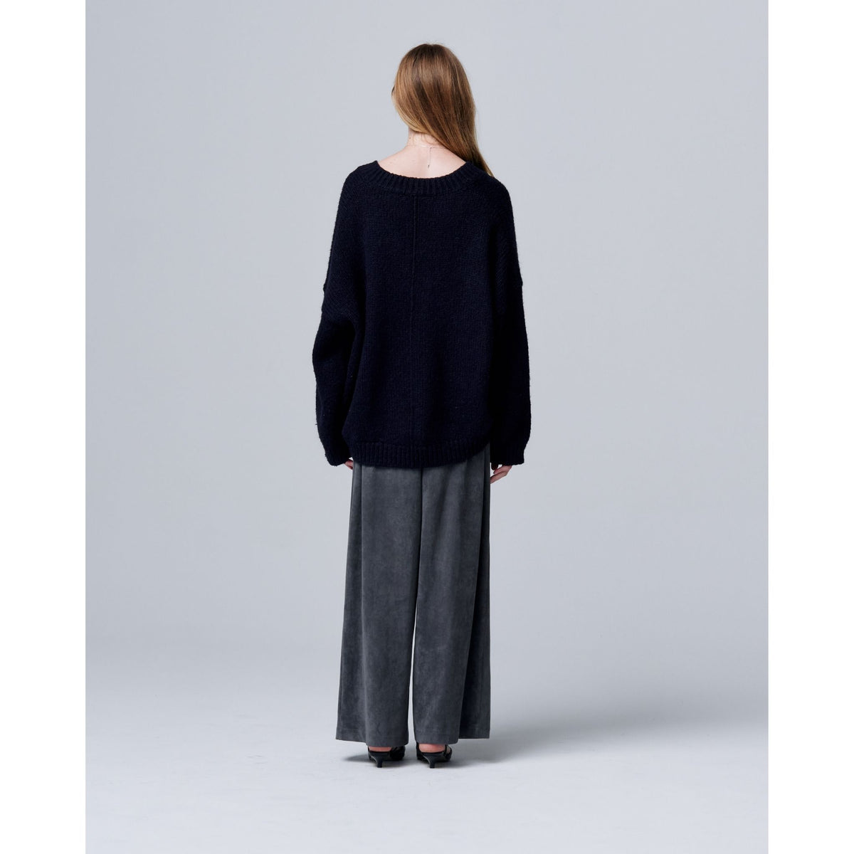 A woman with long brown hair stands with her back to the camera, wearing ODDPRESENT Velour Suede Pants with Elastic Waistband and a loose black sweater, set against a plain light gray background.