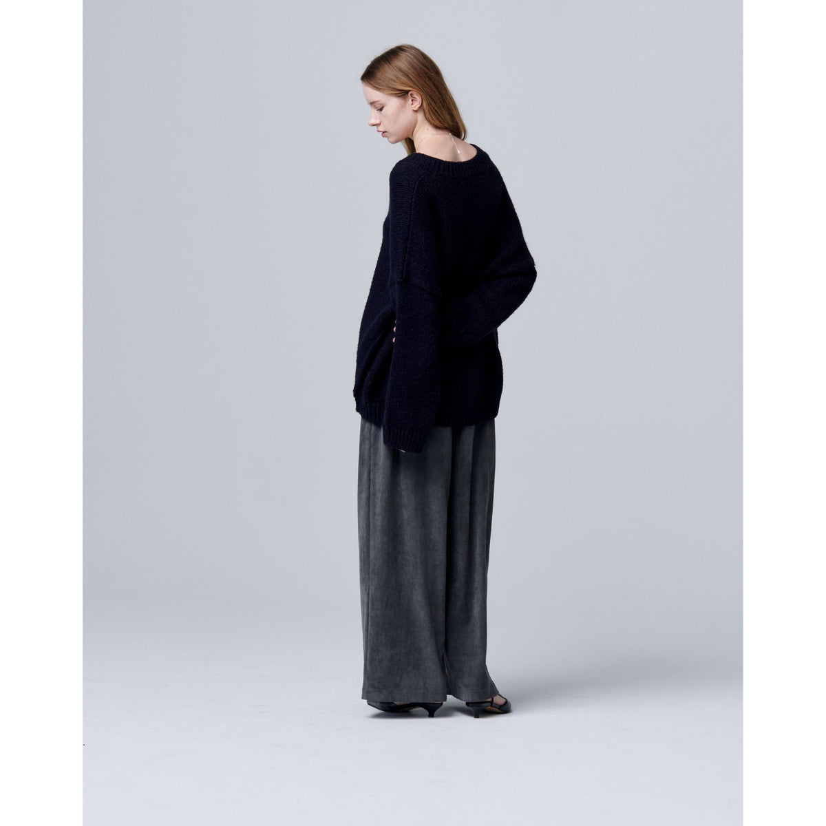A woman stands in a minimal, light gray studio, facing away. She wears ODDPRESENT Velour Suede Pants with Elastic Waistband—long, wide-leg style—paired with a loose black sweater and black shoes.