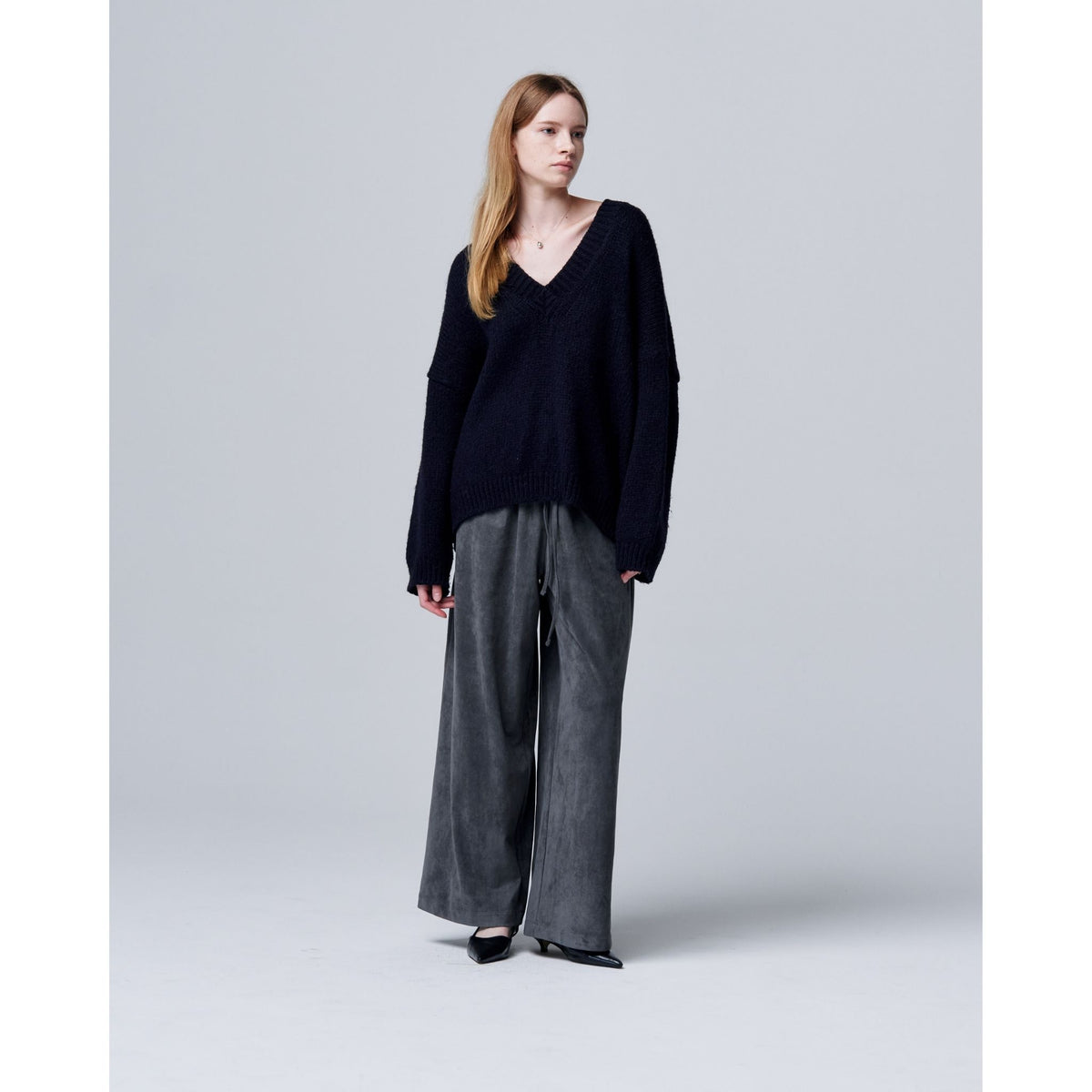 A woman with long, straight hair and a neutral expression stands against a gray background wearing ODDPRESENT Velour Suede Pants with Elastic Waistband, a loose black V-neck sweater, and black pointed shoes.