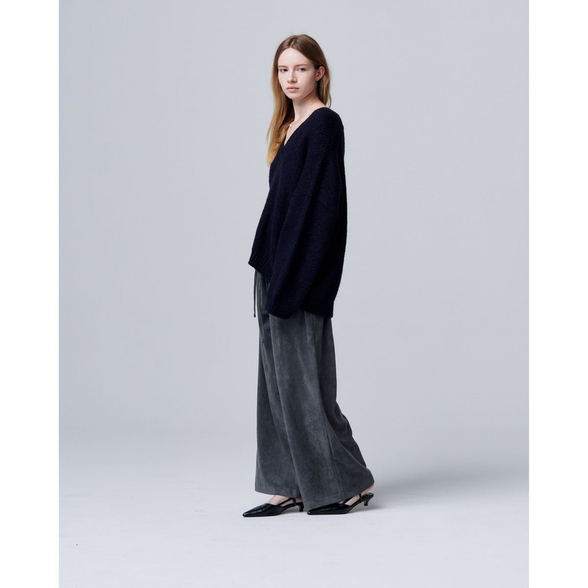 A woman with long straight hair and a neutral expression stands sideways against a light gray background, wearing ODDPRESENT Velour Suede Pants with Elastic Waistband, a loose black sweater, and black shoes.