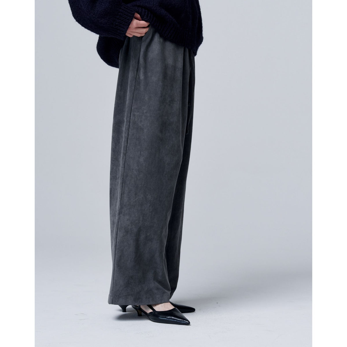 A person in ODDPRESENT Velour Suede Pants with Elastic Waistband, a dark sweater, and black pointed-toe heels stands against a plain light gray background.