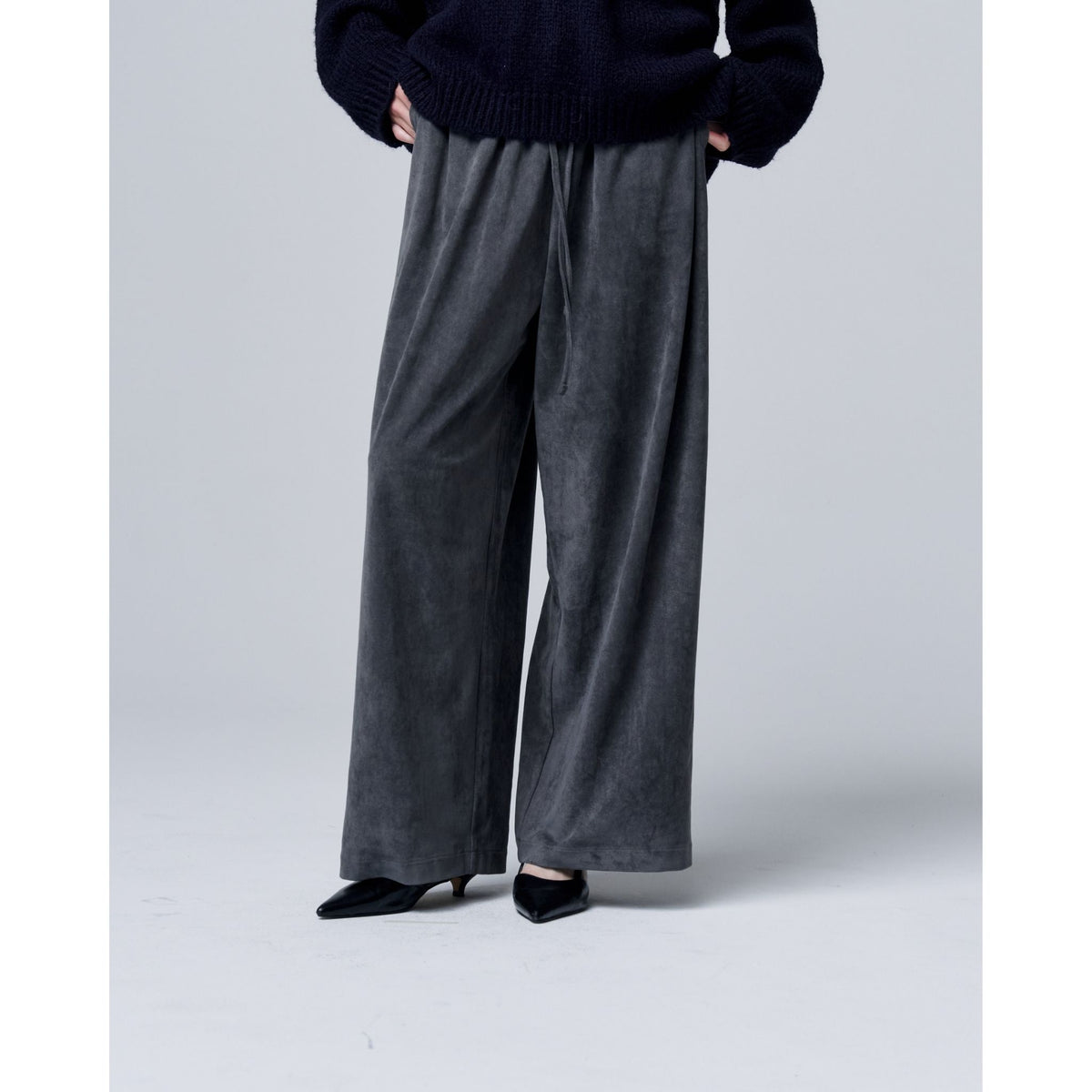 A person models ODDPRESENT Velour Suede Pants with Elastic Waistband—loose-fitting and gray—paired with a dark oversized sweater and black pointed-toe heels. They stand against a plain light background; image cropped at the shoulders.