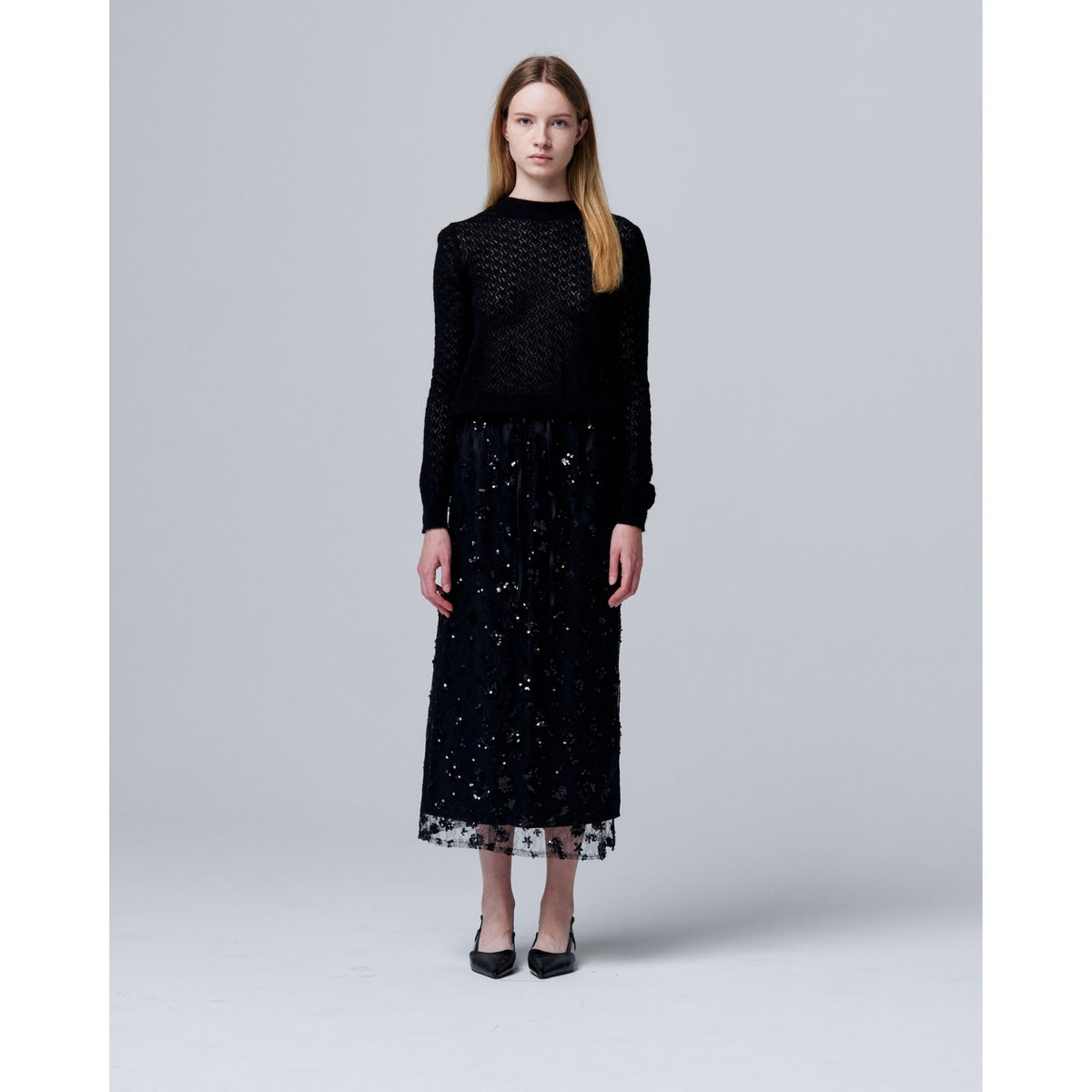 Against a plain gray background, a woman with straight light brown hair models ODDPRESENT’s Sequin Beaded Skirt with Floral Detailing, paired with a black long-sleeve top and black pointed-toe heels—an elegant evening look.
