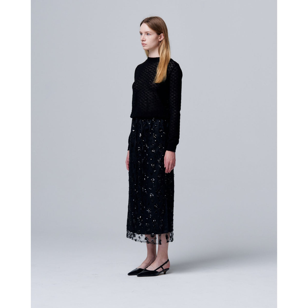 A woman with long light brown hair stands in profile against a gray background, wearing a black top, black flats, and the ODDPRESENT Sequin Beaded Skirt with Floral Detailing featuring an adjustable waist—ideal for evening events.