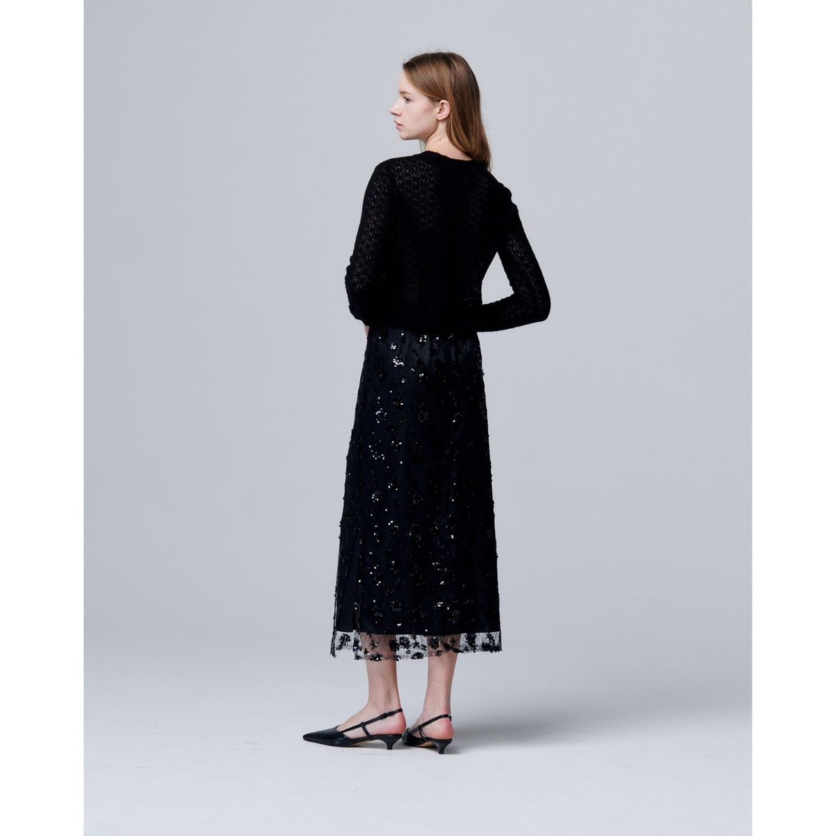 A woman with straight light brown hair faces away, wearing a black textured long-sleeve top, black pointed-toe flats, and the ODDPRESENT Sequin Beaded Skirt with Floral Detailing against a plain light gray background.