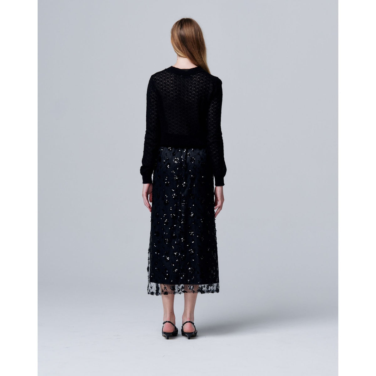 A woman with long, straight hair faces away, wearing a black textured sweater and the ODDPRESENT Sequin Beaded Skirt with Floral Detailing, featuring an adjustable waist—ideal for evening events. She pairs it with black shoes against a plain gray background.