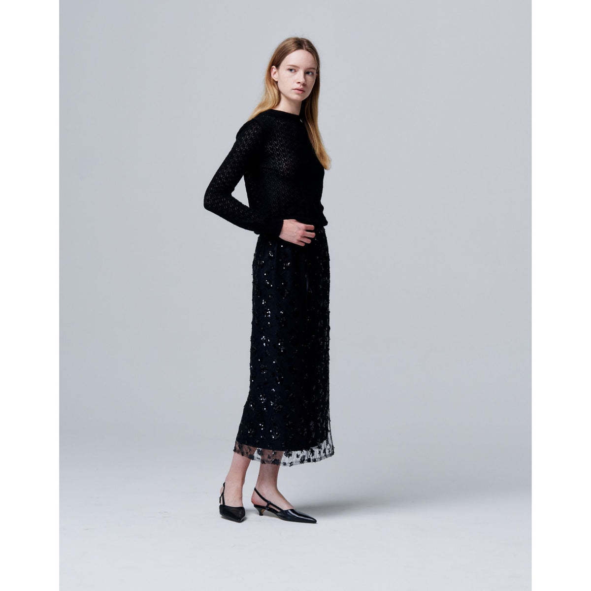 A woman with long blonde hair stands against a light gray background, wearing a black long-sleeve top and the ODDPRESENT Sequin Beaded Skirt with Floral Detailing—a chic choice for evening events.