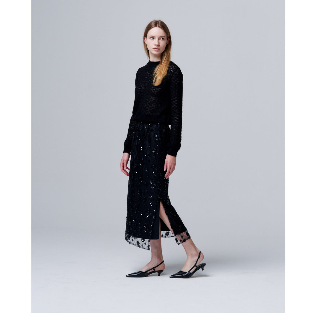 A woman with straight light brown hair and a neutral expression stands against a plain background, wearing an ODDPRESENT Sequin Beaded Skirt with Floral Detailing, a black textured long-sleeve top, and black slingback heels.