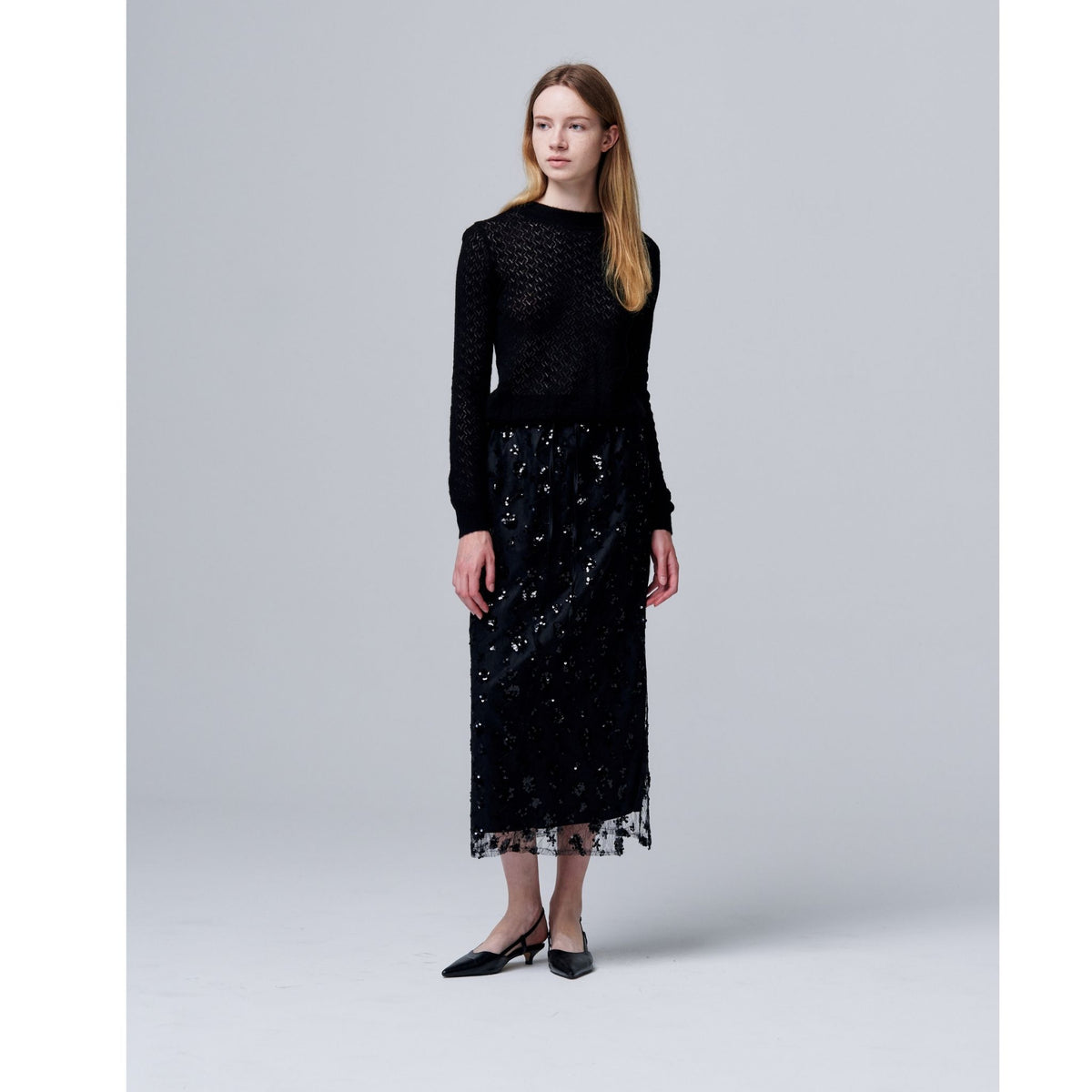 A woman stands against a light gray background wearing ODDPRESENT’s Sequin Beaded Skirt with Floral Detailing, a black long-sleeve knit top, and black pointed-toe heels—an elegant look for evening events. She gazes to the side with a neutral expression.