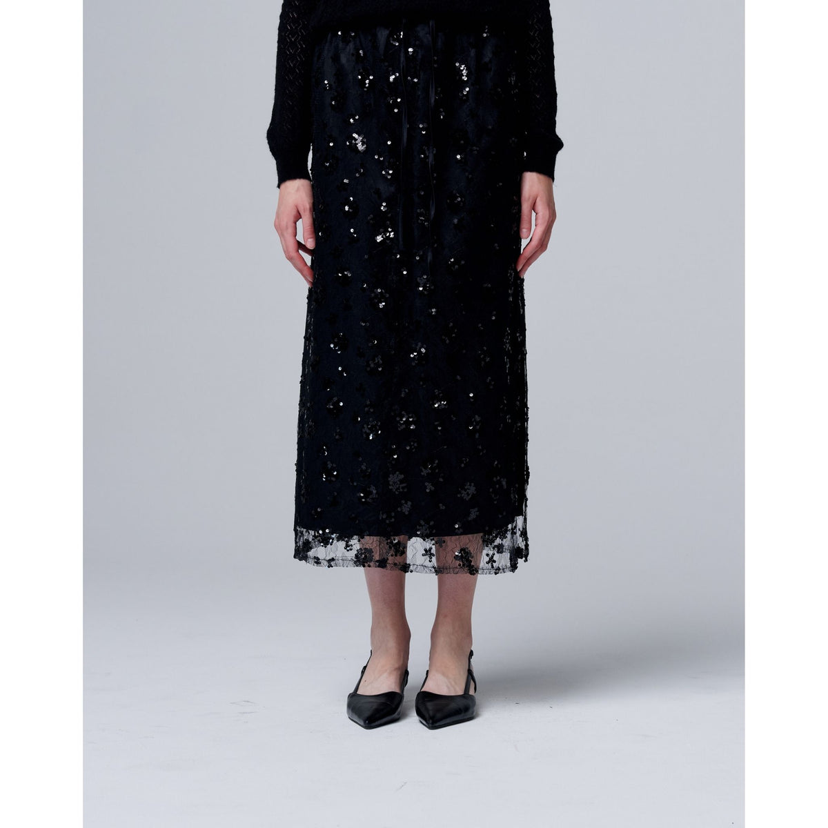 A person wears an ODDPRESENT Sequin Beaded Skirt with Floral Detailing, featuring an adjustable waist strap, paired with a black long-sleeve top and pointed-toe heels—perfect for evening events—while standing against a plain light background.