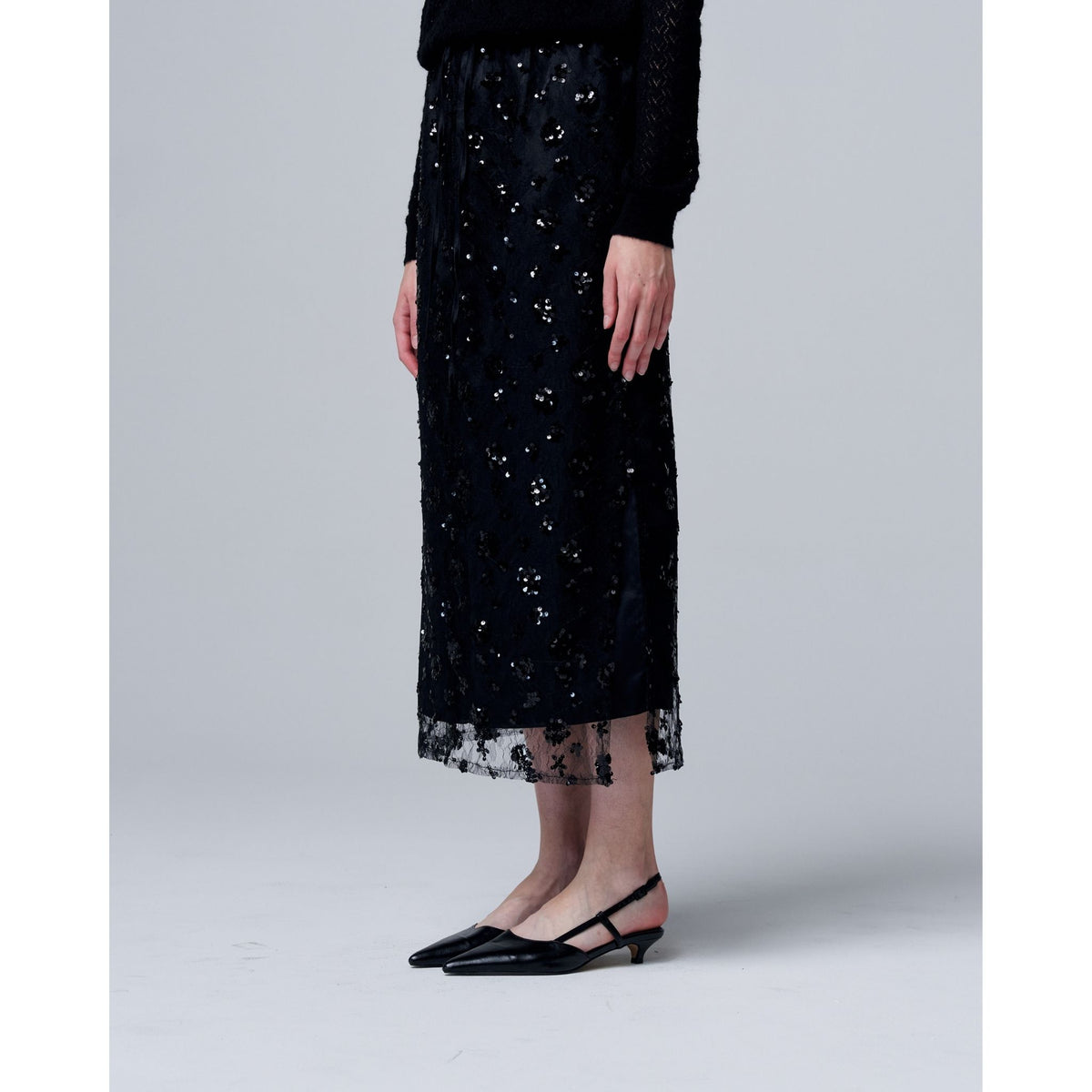 Dressed in a black long-sleeve top, the ODDPRESENT Sequin Beaded Skirt with Floral Detailing, and black pointed-toe slingbacks against a light background—an elegant look for evening events.