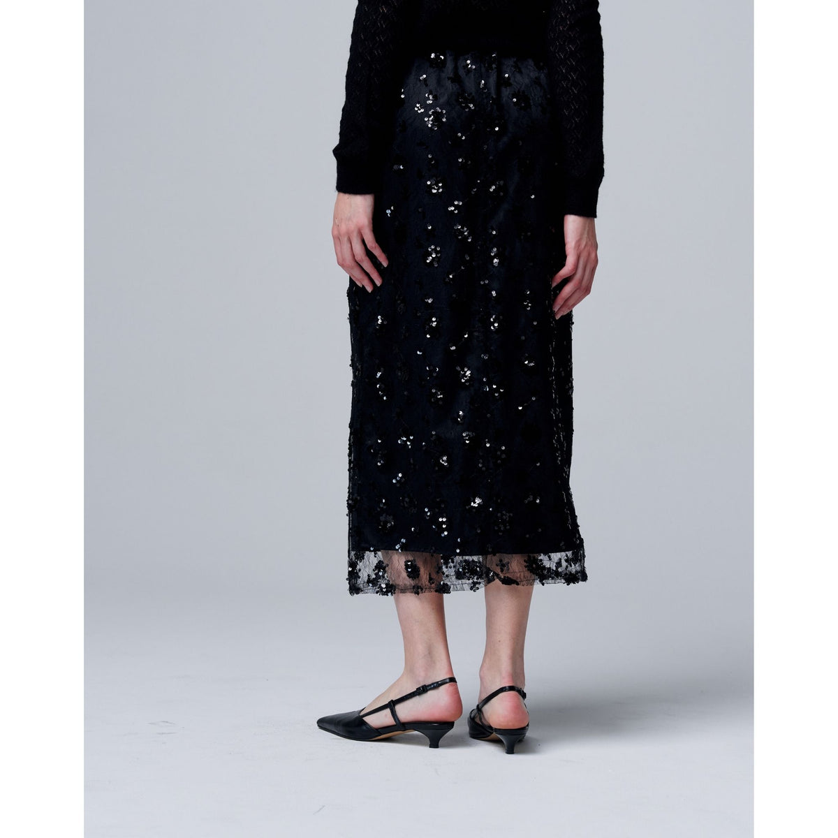 A person wears the ODDPRESENT Sequin Beaded Skirt with Floral Detailing, featuring an adjustable waist strap, paired with black T-strap low heels against a light gray background—ideal for evening events.